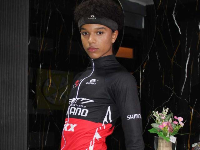 LiamMiranda with curly black hair, black headband, and black-red cycling jersey, standing with a serious face, holding a bouquet of pink flowers. Black marble background.