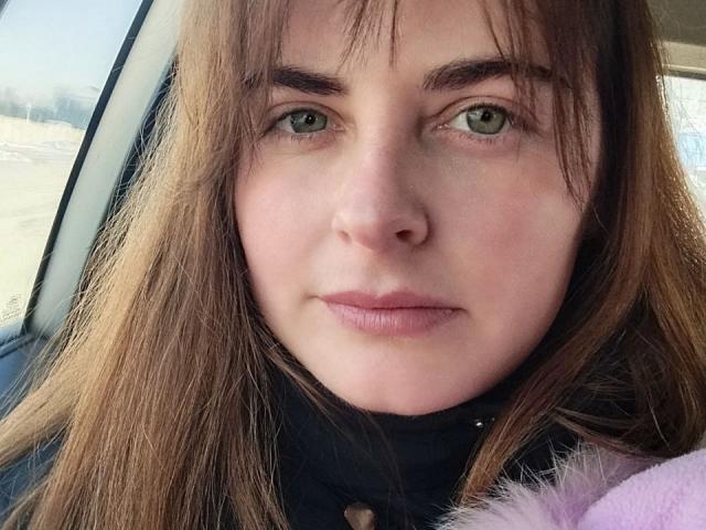 Angela Winter tem olhos verdes, cabelo castanho e uma expressão neutra. Ela está vestindo uma blusa preta com botões e tem um objeto rosa e felpudo em primeiro plano..