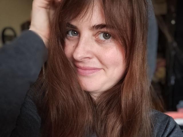 Angela Winter, com cabelo castanho escuro, olhos azuis e um leve sorriso, vestindo um suéter cinza escuro, tocando a cabeça com a mão. Seus seios não estão visíveis..