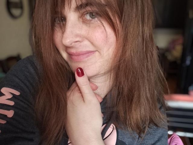 Angela Winter mit braunen Haaren, roten Nägeln, die leicht lächelt, sich an ihr Kinn fasst, ein graues Hemd trägt, ein süßes Gesicht zeigt und einen hellen Hautton hat..