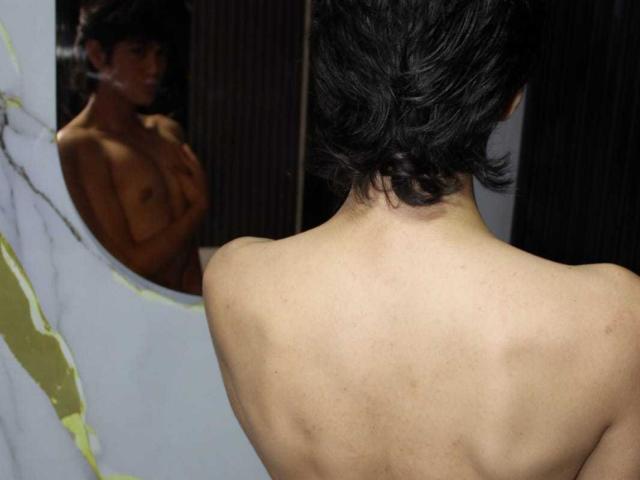MarcoMiranda, shirtless, facing mirror, back visible, short black hair, brown skin, muscular, reflecting in mirror, bare chest with small, dark nipples, looking down, dark room.