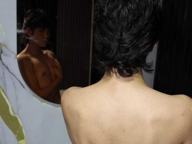 MarcoMiranda with short black hair, seen from behind, looking at his reflection in a mirror, showing his bare back and small, brown, tattooed tits.