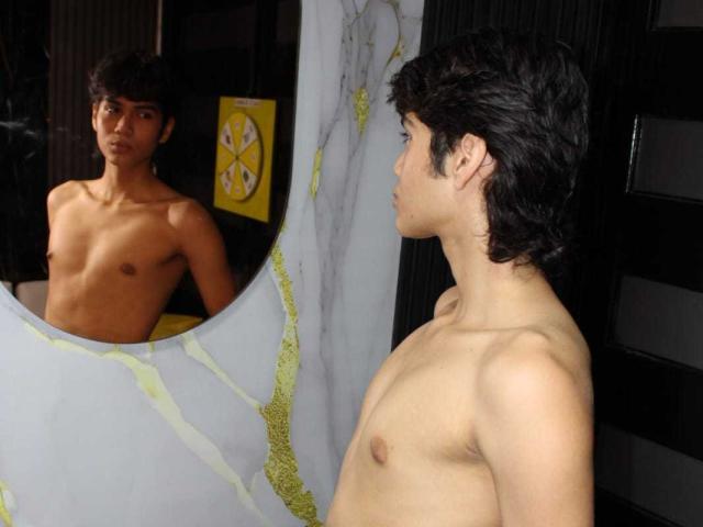 MarcoMiranda with a lean, shirtless build, dark hair, and small tits, stands in profile, staring at his reflection in a mirror, showing his bare chest and nipples.