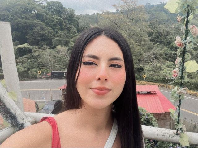 EimmyPark tem cabelos pretos longos, blush rosa e delineador preto, veste uma blusa vermelha, sorri com lábios rosados e está em pé ao lado de um poste branco com decorações florais..