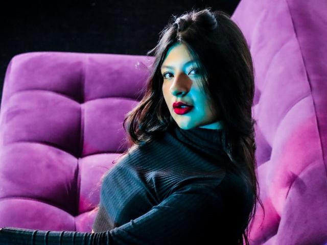 ClaraPattel mit rotem Lippenstift, langen, dunklen Haaren, einem schwarzen, gerippten Rollkragenpullover, sitzt auf einem violetten, gepolsterten Sofa, blickt über die Schulter, blaues Licht auf der rechten Seite, rosa Licht auf der linken Seite..