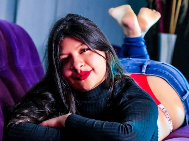 ClaraPattel está acostada en un sofá morado, con las piernas levantadas, vistiendo ropa interior roja y un jersey negro de cuello alto, con el pelo largo y negro y pintalabios rojo, sonriendo. Sus pies están en el aire..