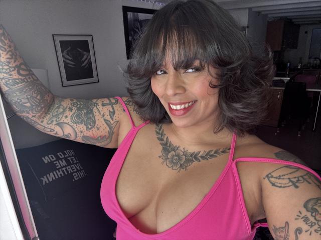 Chili com cabelo encaracolado escuro, vestido rosa, tatuagens nos braços e no peito, sorrindo, decote visível, braço levantado, dentes sorridentes.