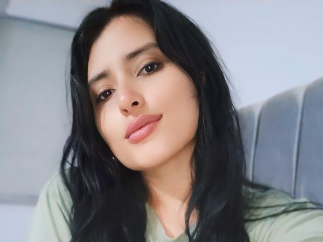 VaneHot69 heeft lang zwart haar, een lichtbruine huid en volle, roze lippen. Ze draagt een groene blouse en kijkt iets omlaag, met een subtiele glimlach..