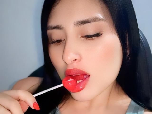 VaneHot69 com cabelo preto, batom vermelho e língua rosa brilhante sugando uma pirulito vermelha, olhos semicerrados, segurando-o com dedos com unhas vermelhas, em uma foto semelhante a um selfie..