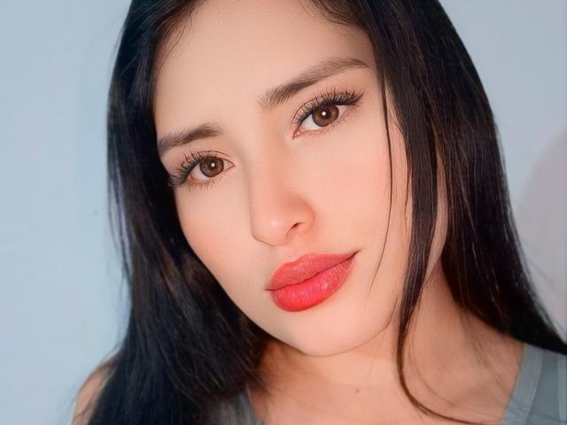 VaneHot69 hat langes, schwarzes Haar, braune Augen und roten Lippenstift. Ihr Gesicht ist zentriert und zeigt ein leichtes Lächeln. Sie trägt ein graues Oberteil und hat einen hellen, zarten Hautton..