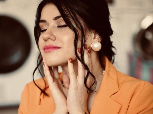 Anyaxxs with closed eyes, pink lipstick, and orange blazer, touching her face with red-manicured fingers, wearing large white earbuds, has wavy black hair and a calm expression.