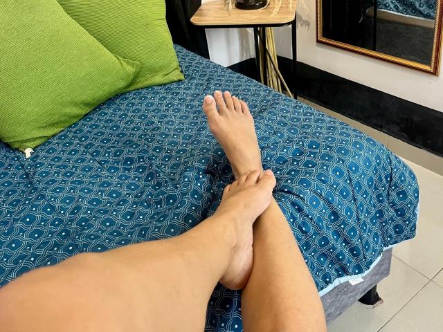 DaliaJohnson's legs are crossed at the ankles, showing her bare feet on a blue patterned bedspread with green pillows. Her skin is tan, and the bed is in a room.