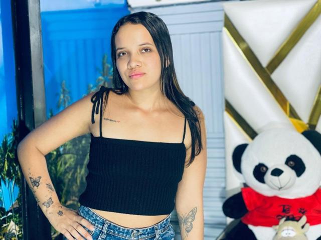 NathashaWilt, avec de longs cheveux bruns, un débardeur noir côtelé, un short en jean, des tatouages sur les bras, debout, les mains sur les hanches, mignonne, petite poitrine, et un ours en peluche panda en arrière-plan..