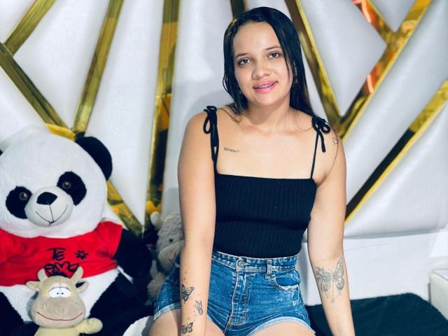 NathashaWilt, portant un débardeur noir qui révèle de petites seins, et un short en jean, assise avec des tatouages et des pandas en peluche, souriant. Elle a des hanches de taille moyenne, des cheveux foncés et une peau claire..