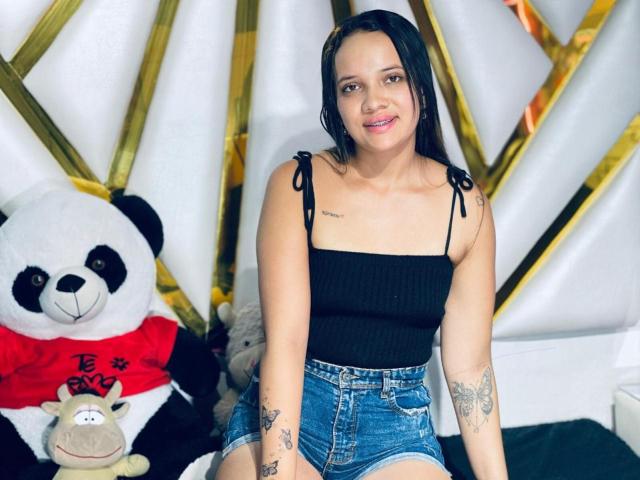 NathashaWilt, portant un débardeur noir qui révèle de petites seins, des bras tatoués et des yeux marron, assise devant un ours en peluche géant, vêtue de shorts en jean..
