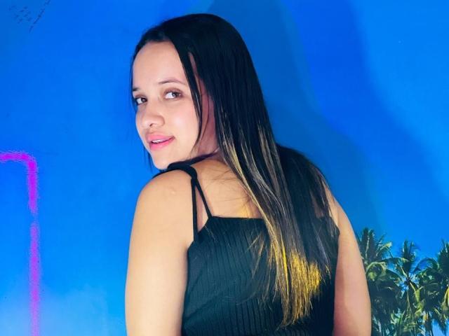 NathashaWilt, avec de longs cheveux noirs, portant un haut noir sans manches, regardant par-dessus son épaule, souriant avec des lèvres roses, fond bleu, et une plante verte sur la droite..