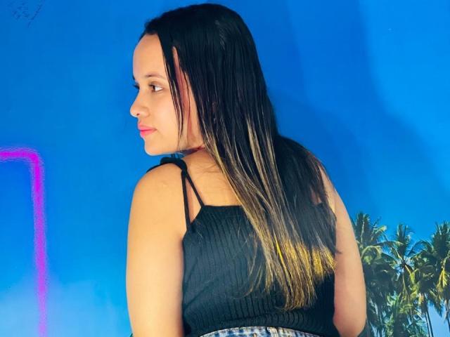 NathashaWilt met lang zwart en blond haar in een ombre-stijl, draagt een zwart ribtrui en kijkt over haar schouder, waarbij haar rug en een licht gedraaid profiel zichtbaar zijn..