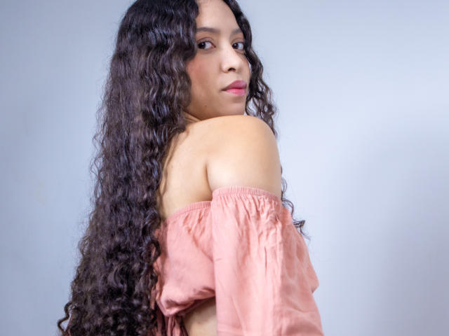 ClarelandsWell with long, curly dark hair, pink off-shoulder top, looking back, showing bare shoulder, soft expression, light skin, subtle makeup, casual style, simple background.