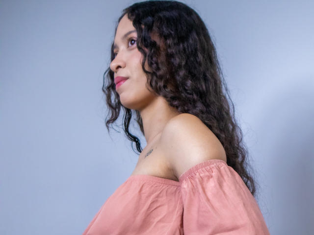 ClarelandsWell with curly dark hair, pink off-shoulder top, and small tattoo on her right shoulder, smiling sideways, showing her profile and slight breast.