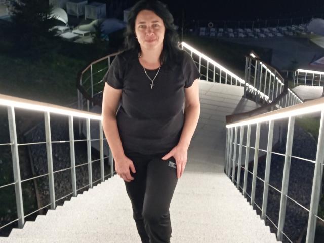 MuseOfSelene stands on a metal staircase at night, wearing a black shirt, black pants, and a cross necklace, smiling with hands on her thighs, dark wavy hair.