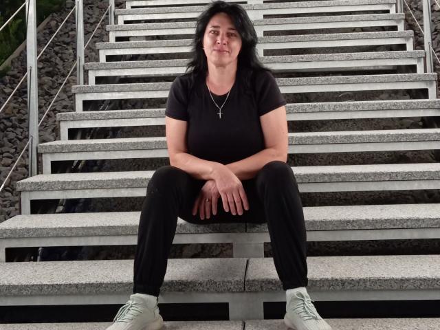 MuseOfSelene is a middle-aged woman with black hair, black shirt, black pants, and white sneakers, sitting on gray concrete stairs with her hands on her knees.