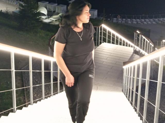 MuseOfSelene, wearing a black t-shirt and black pants, has a cross necklace, walking up a lit staircase at night, with a slight smile, holding a backpack.