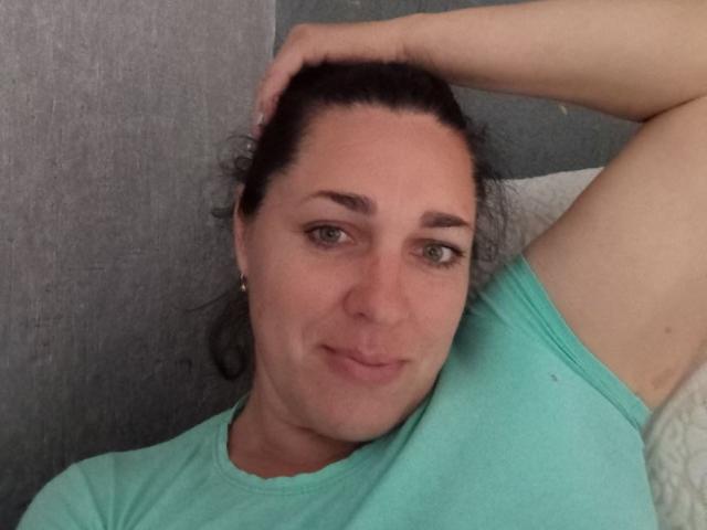 MuseOfSelene has puffy tits, green eyes, and a slight smirk. She's wearing a teal shirt, lying on a gray surface with her arm behind her head.