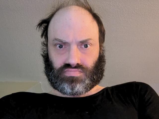 Un homme chauve, avec une barbe, portant des collants et une chemise noire, avec des sourcils gris, regardant droit devant la caméra avec une expression neutre..