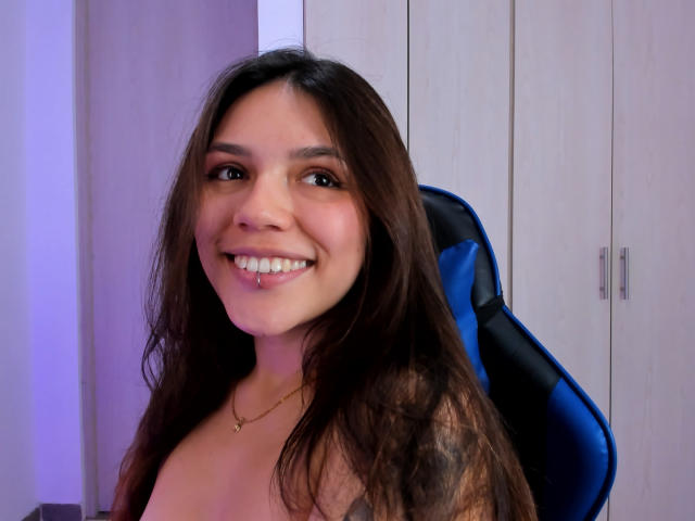 SophiaMarx with long brown hair, smiling, sitting in a blue chair, wearing a necklace, showing her tits, has a tattoo on her right arm, and cute pink lips.