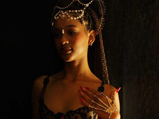 Ally Mills, con la pelle scura, i capelli intrecciati, una corona di perle, un abito di pizzo nero, le unghie rosse, un anello d'oro e un sorriso sicuro, illuminata da una luce soffusa e calda. Il seno è visibile, appare sexy..