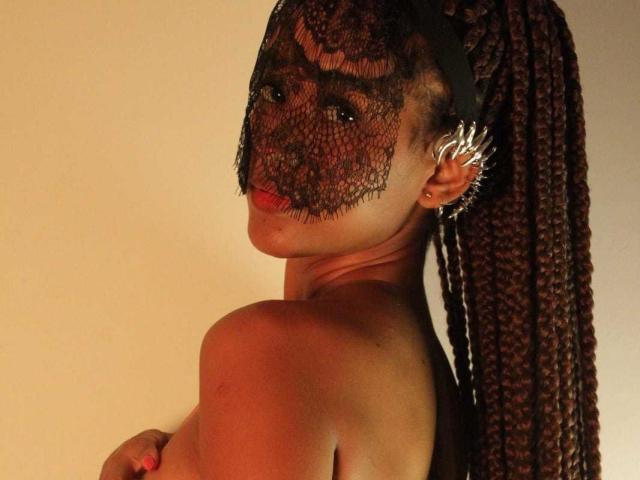 Ally Mills mit langen, braunen Zöpfen, die eine schwarze Spitzenmaske trägt, glänzende Ohrringe, roten Lippenstift und die über die Schulter schaut, mit freiem Dekolleté und einem sexy, selbstbewussten Ausdruck..