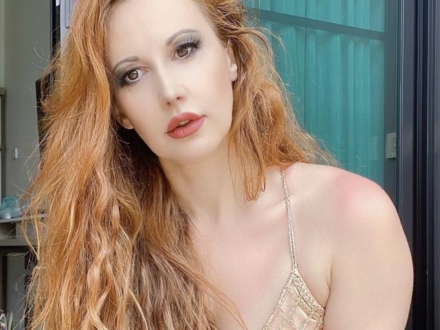 MayaGloss has wavy, dirty-blonde hair, light skin, and brown eyes. She's wearing a lace, beige top with exposed shoulders. Her lips are slightly parted and she's looking at the camera.
