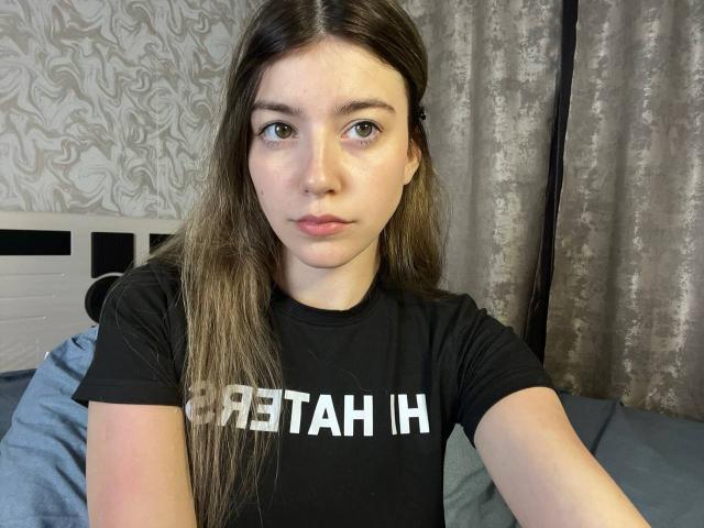 VinnaSkyHott met lang bruin haar, lichte huid en blauwe ogen, gekleed in een zwart T-shirt, zittend op een bed met een neutrale uitdrukking, waarbij haar kleine borsten en volle lippen zichtbaar zijn..
