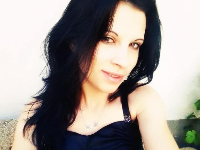 ClaraSweetty with black hair, brown eyes, and a slight smile, wearing a black bra, showing her tits, and a delicate necklace against a white background.