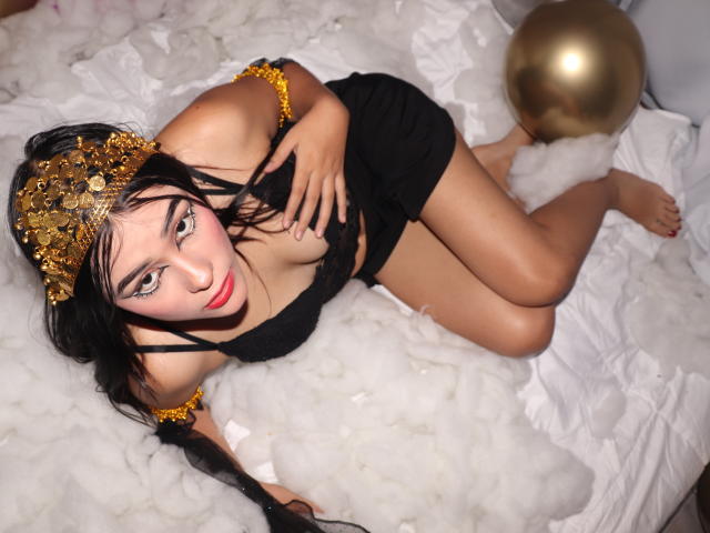 JulietaGarcia with black hair, gold headpiece, and black outfit, lying on white fluffy stuff, gold ball in background, looking up with red lips, touching her tits.