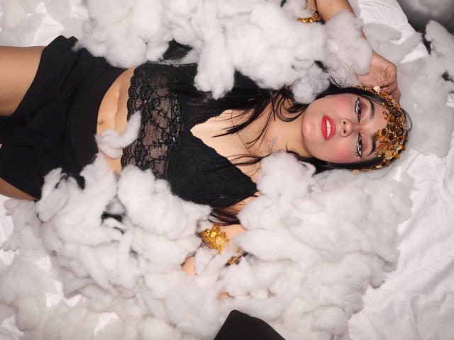 JulietaGarcia with black lace top, red lips, gold headpiece, lying in fluffy white smoke, wearing black skirt, gold bracelets, her tits visible, arms above head, looking at camera.