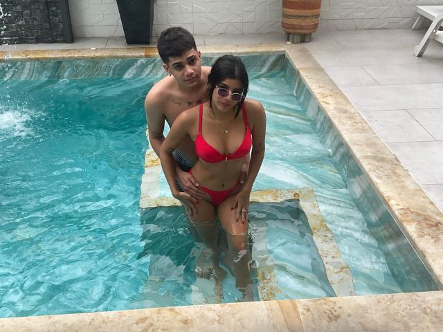 RileyAndChriss, avec un bikini rouge, ses seins visibles, debout dans une piscine, Chriss derrière elle, les mains sur ses fesses, toutes les deux portant des lunettes de soleil, l'eau leur arrivant aux cuisses..