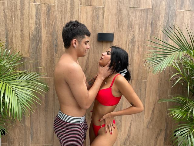 RileyAndChriss, torse nu, avec un petit pénis circoncis, tient le menton d'une femme noire, aux cheveux noirs, de taille moyenne, vêtue d'un soutien-gorge et d'une culotte rouges. Elle embrasse son cou, et il y a des plantes vertes de chaque côté..