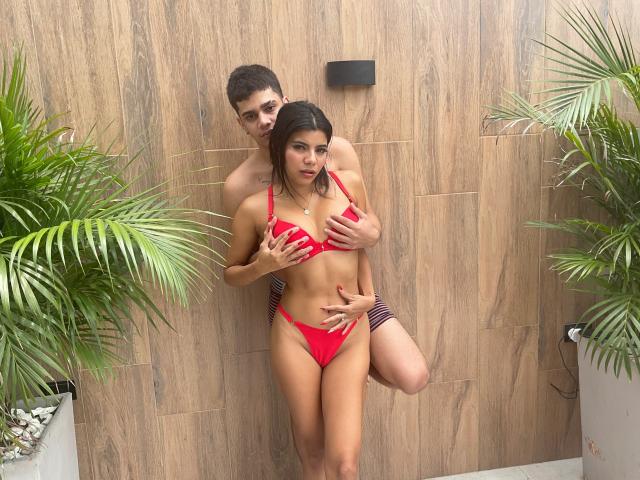 RileyAndChriss em lingerie vermelha, seus seios cobertos pelas mãos dele, em pé contra uma parede de madeira, com plantas de palmeira verdes nos lados. Chriss está atrás, com as mãos nos quadris e nos seios..