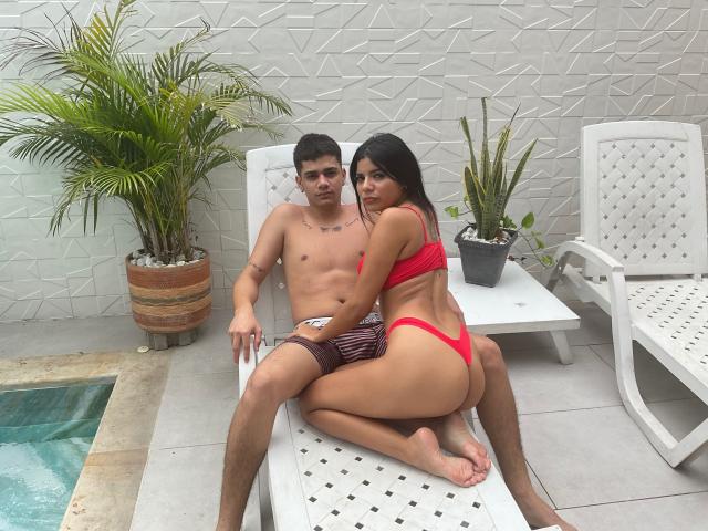 RileyAndChriss, ohne Hemd und mit einem kleinen Tattoo auf der Brust, sitzt auf einem weißen Liegestuhl. Chriss, in einem roten Bikini, kniet und zeigt ihr Gesäß, während er seine gestreiften Shorts trägt..