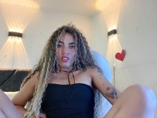 LiliRose69 with long, curly blonde hair, black strapless top, tattoo on right arm, legs spread, sitting on bed, looking at camera with slightly open mouth.