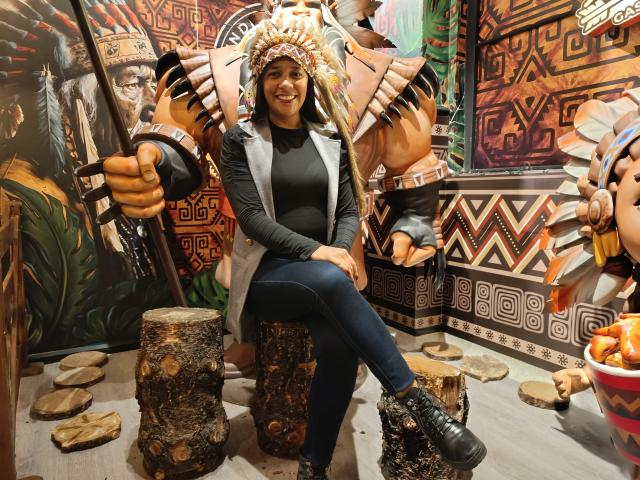 PauletteLoveX sitting with crossed legs, wearing a Native American headdress, black long-sleeve top, blue jeans, and black boots, surrounded by wooden stools and decorative patterns.
