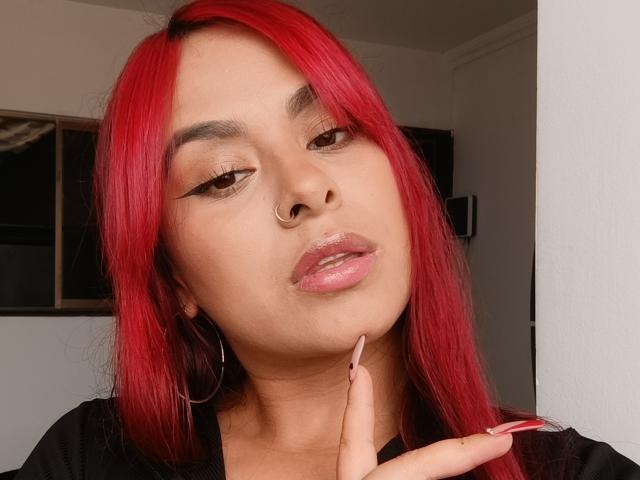 EmilyEvanss with red hair, nose ring, and shiny pink lips, looking down with finger on chin, wearing black shirt, hoop earrings, and long pink nails.