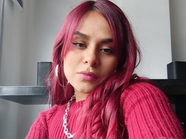 EmilyEvanss with pink hair, wearing a red ribbed sweater, hoop earrings, and a silver chain, has a nose ring and full pink lips, looking slightly annoyed.