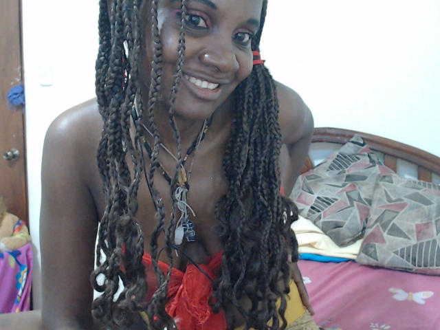 Katrhina87-hot with dark brown skin, long braided hair, smiling, wearing red lace lingerie, showing her tits, sitting on a bed with patterned pillows and a wooden headboard.