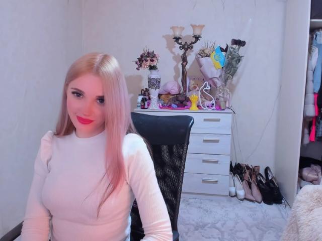 AlienNina with pink hair, white long-sleeve top, red lipstick, smiling, sitting in black chair, white drawers, candelabrum, flowers, and shoes in background. Boobs, cute, pink hair.