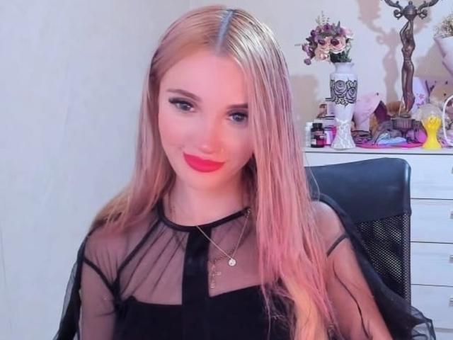 AlienNina with long blonde hair, red lipstick, black sheer top, small tits, necklace, sitting in black chair, smiling, pink and purple decor in background.