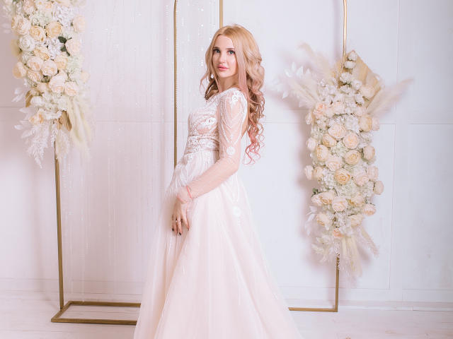 AlienNina with pink wavy hair in a sheer lace, long-sleeve, white wedding dress, standing in front of floral arrangements. Her tits are visible through the lace.