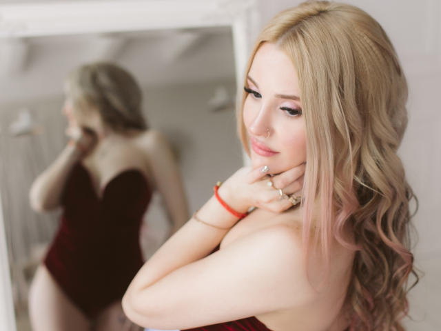 AlienNina with blonde hair, wearing a red velvet bodysuit, looking at herself in a mirror, showing her tits and a small nose ring, with a red bracelet on.