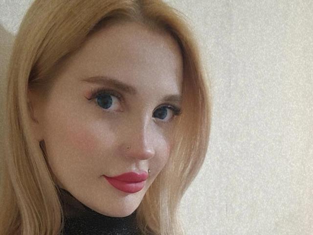 AlienNina has blonde hair, blue eyes, red lipstick, and nose piercings. She's smiling slightly with a black top. Her tits are not visible. The photo is a selfie.